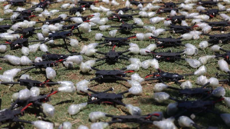 Dozens of black quadcopter drone frames lie scattered on grass alongside numerous bundles wrapped in clear plastic.
