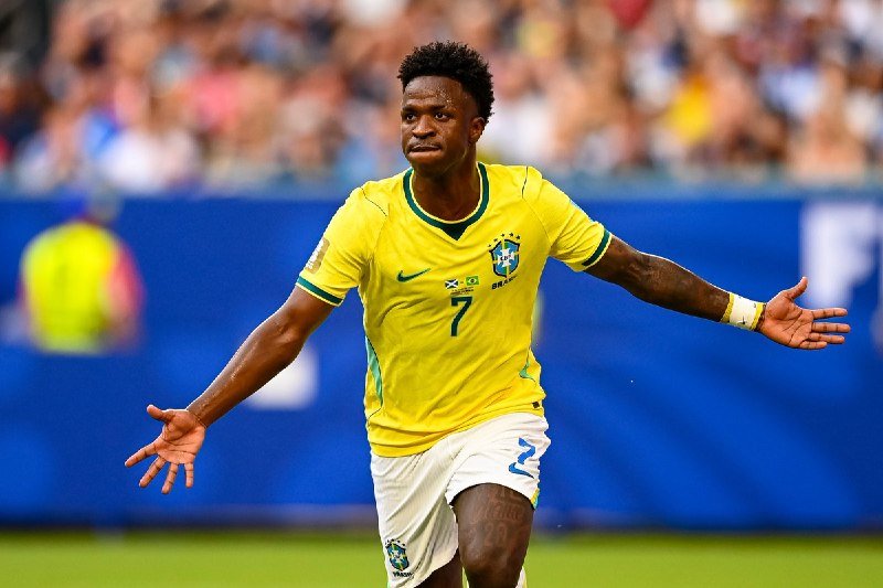 A soccer player wearing a yellow Brazil jersey with the number 7 celebrates with arms outstretched in a stadium filled with spectators.