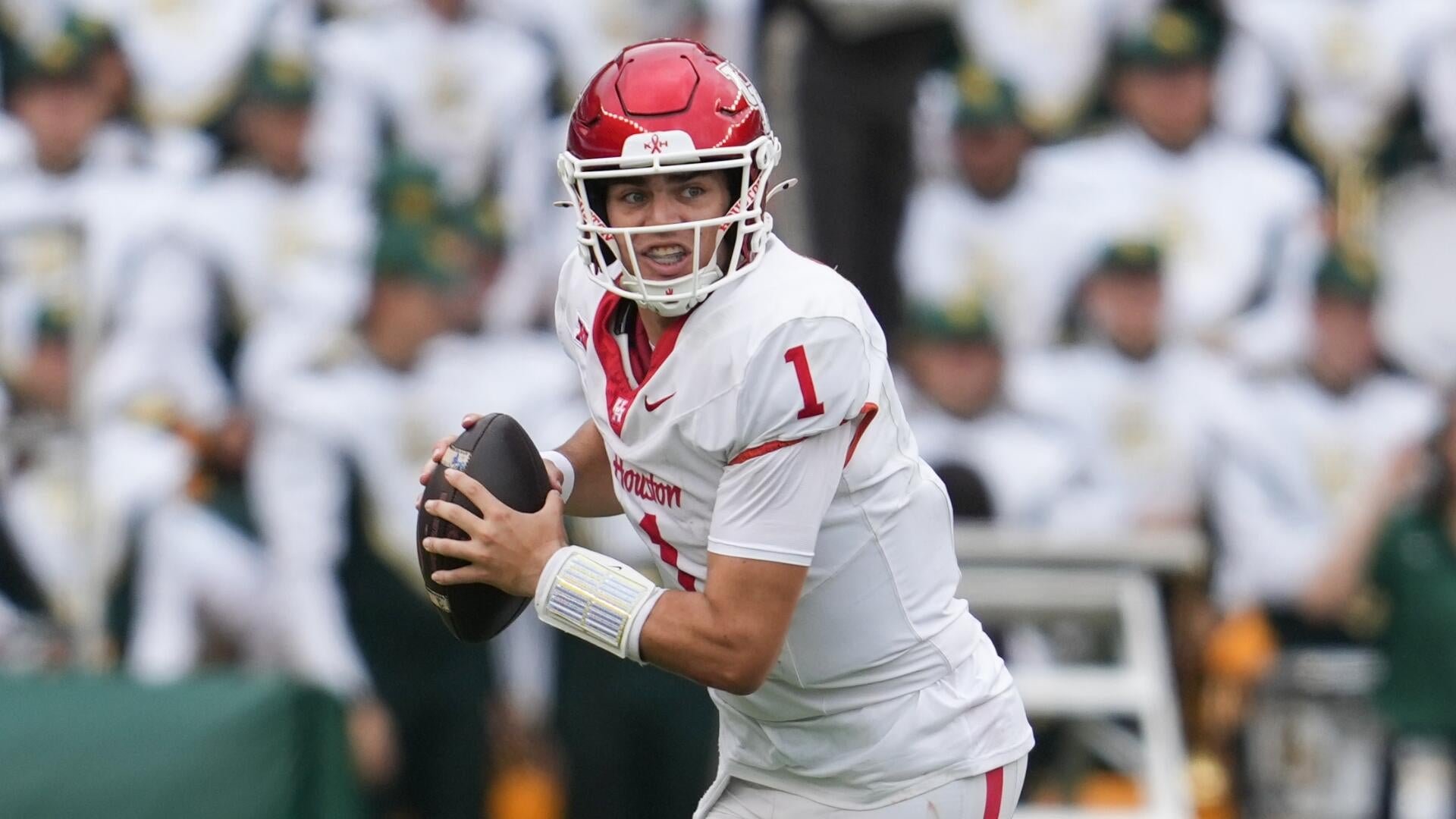 A Houston football quarterback wearing jersey number 1 holds a football while scanning the field, with Baylor players visible on the sideline in the background.