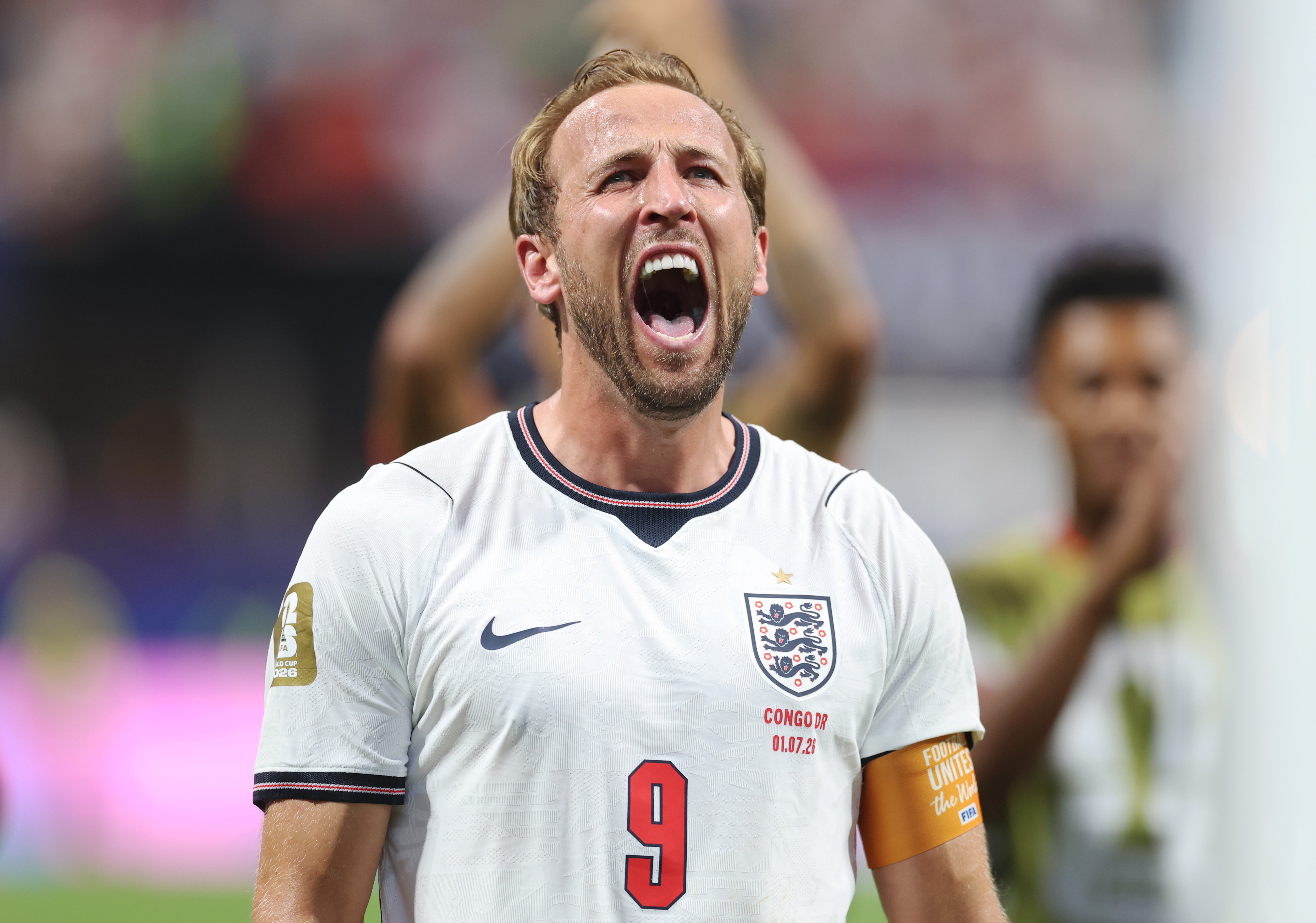 A blonde, bearded soccer player wearing a white England jersey with the number 9 yells passionately during a match, with blurred teammates visible in the background.