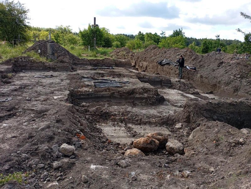 An archaeological excavation site shows a large dug-out pit with exposed stone foundations, surrounded by dirt mounds, with a person working amid green trees.