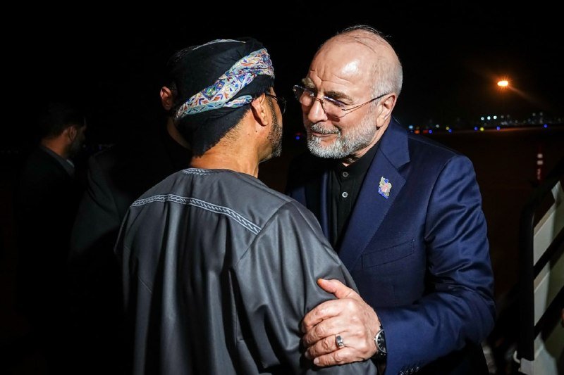 Two men embrace at night on a lit airport tarmac, one wearing a patterned headwrap and gray tunic, the other in a navy suit and glasses.