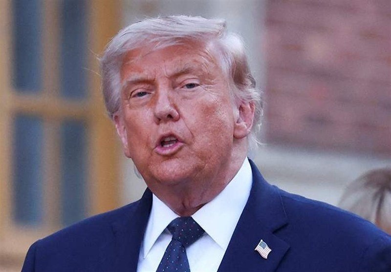 A blonde-haired man in a dark suit and red tie gestures with both hands raised, seated in front of others in an official setting, with Arabic text displayed below.
