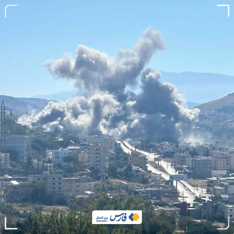 A plume of gray smoke rises above a rural landscape of buildings, trees, and hills in the distance.