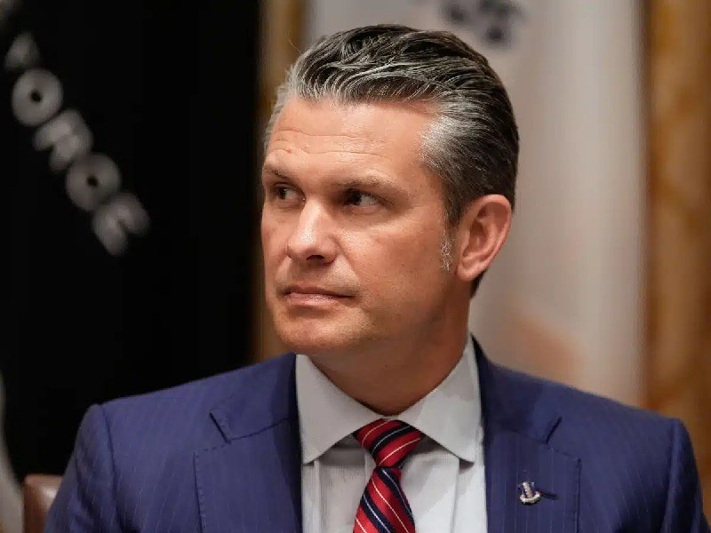 A middle-aged man in a dark suit and tie sits with a serious expression against a blurred wooden background.
