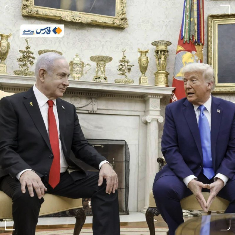 Two men in suits sit facing each other in an ornate room with a fireplace mantel displaying gold trophies, framed paintings, and a military flag.