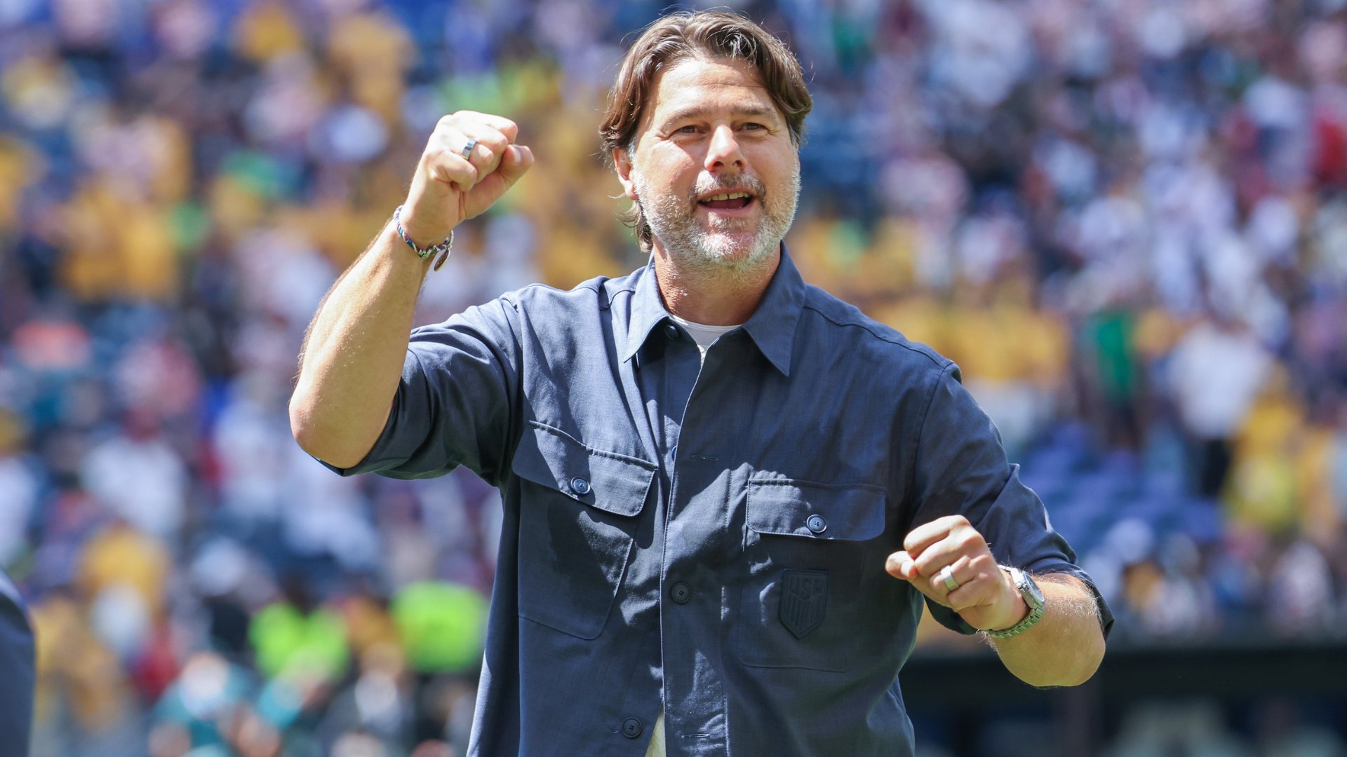 A gray-bearded man in a dark blue shirt raises his fist in celebration, standing before a blurred crowd in a stadium.