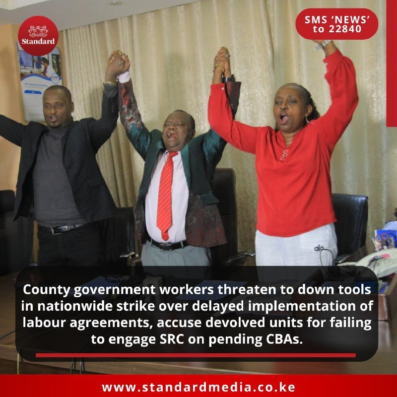 Three people stand with raised arms and clasped hands in an office, appearing to sing or chant.