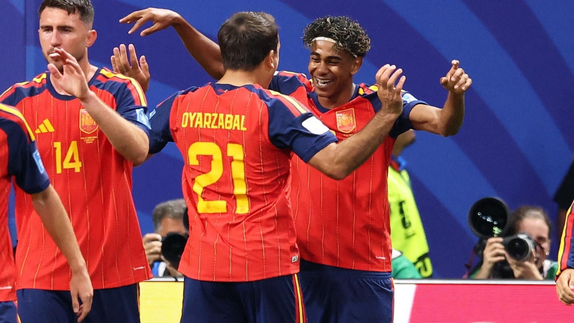 Soccer players in red and yellow jerseys, including one marked "OYARZABAL 21" and another "14," celebrate together on the pitch as photographers capture them.