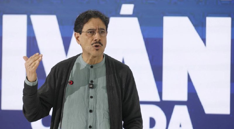 A man with glasses and a mustache, wearing a light green shirt and dark cardigan, gestures with his hand while speaking in front of a large blue screen displaying partial white text.