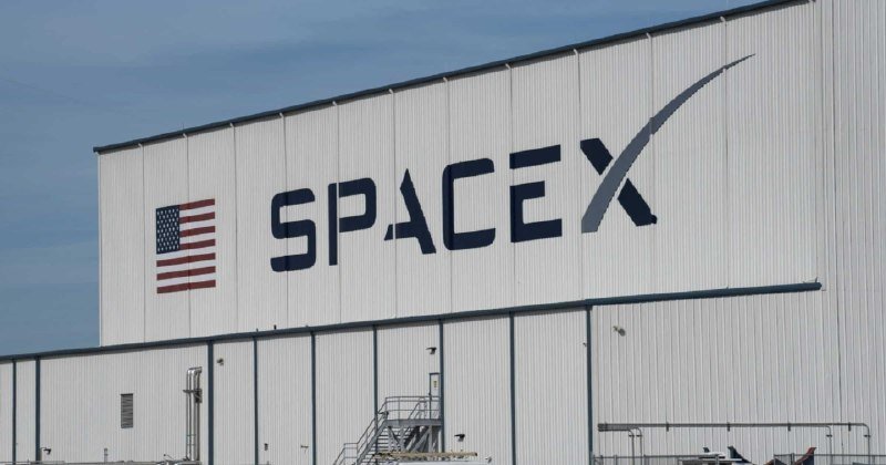 A large white industrial building displays the "SpaceX" logo and an American flag painted on its exterior under a blue sky.