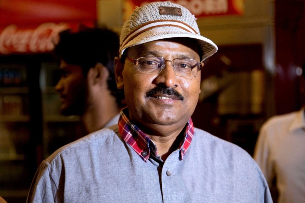 A smiling man wearing glasses, a striped fedora, and a gray shirt over a plaid collar stands in front of a red Coca-Cola sign, with other people visible in the background.