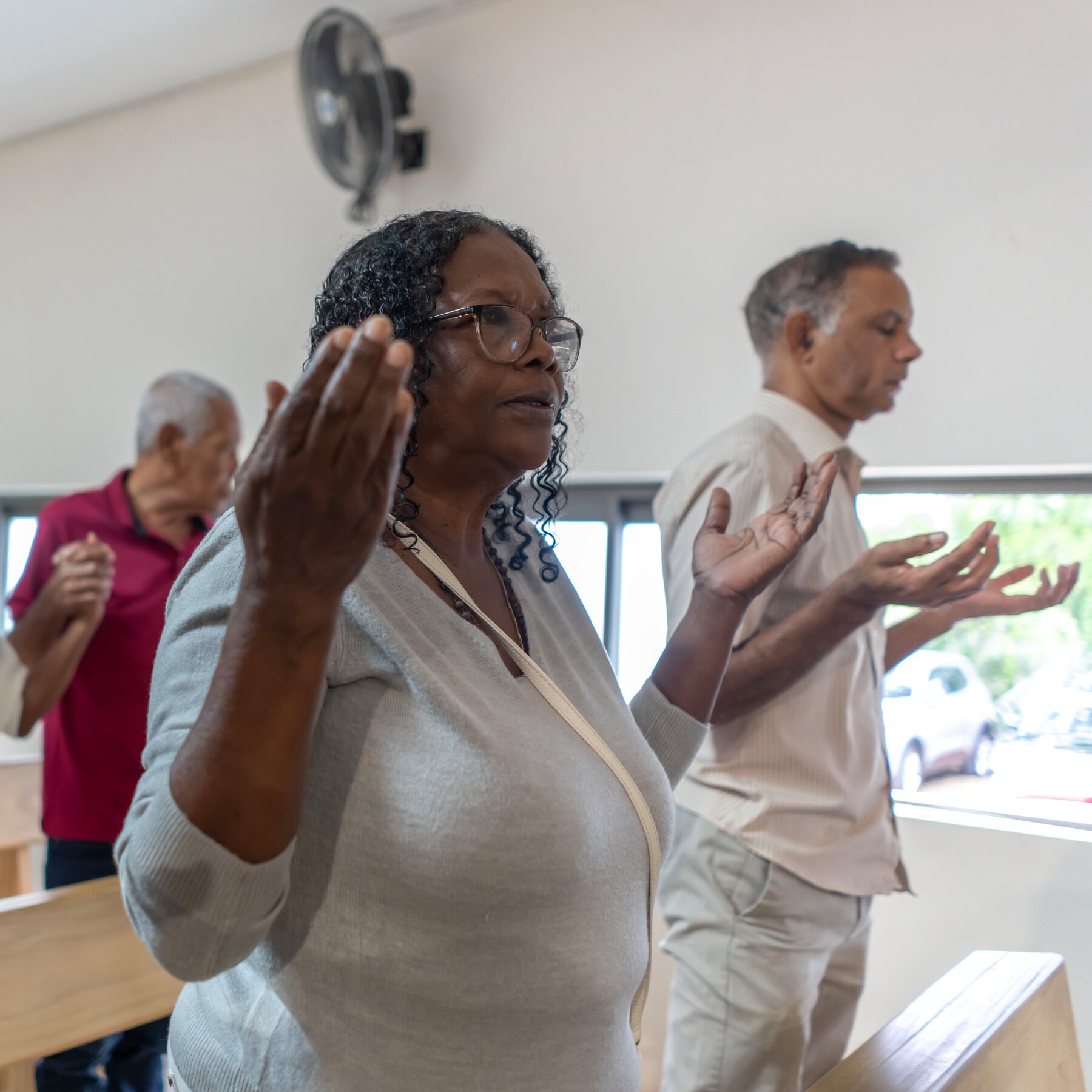 People stand with raised, open hands and closed eyes in an indoor room, appearing to pray or worship.