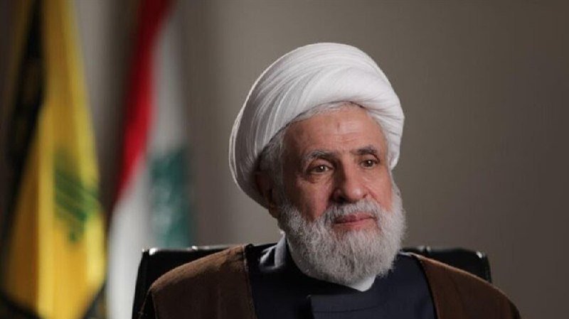 An older man with a white turban and beard sits indoors, with yellow and red-white-green flags visible in the blurred background.
