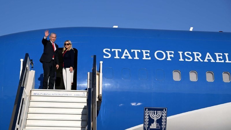 A man in a dark suit waves from the top of airplane stairs next to a woman, standing before a blue aircraft labeled "STATE OF ISRAEL" with the Menorah emblem.