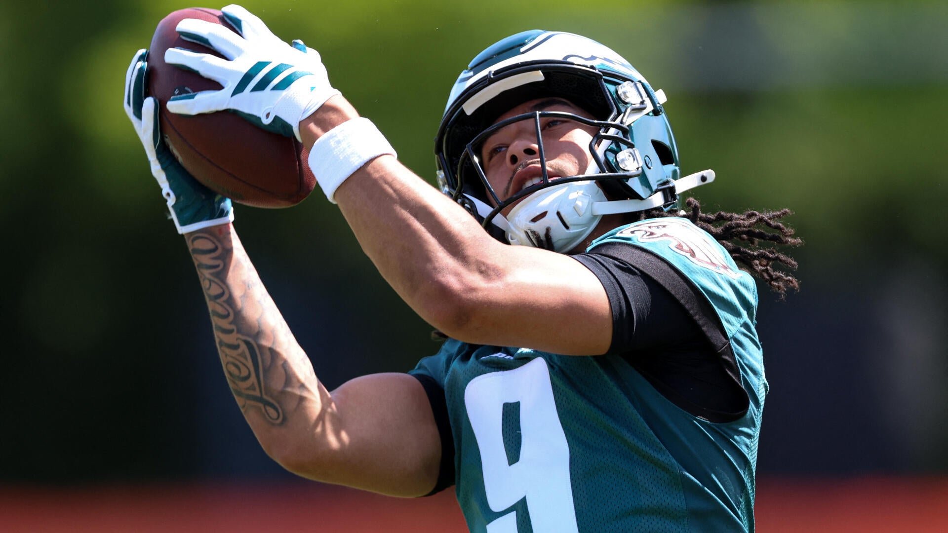A football player wearing a green Philadelphia Eagles practice jersey with the number 9 catches a football with both hands during an outdoor practice session.