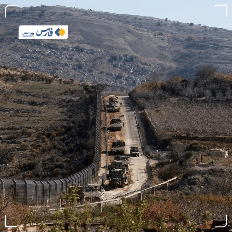 A military convoy of vehicles travels along a fenced border road through a hilly, arid landscape, viewed from an elevated vantage point.