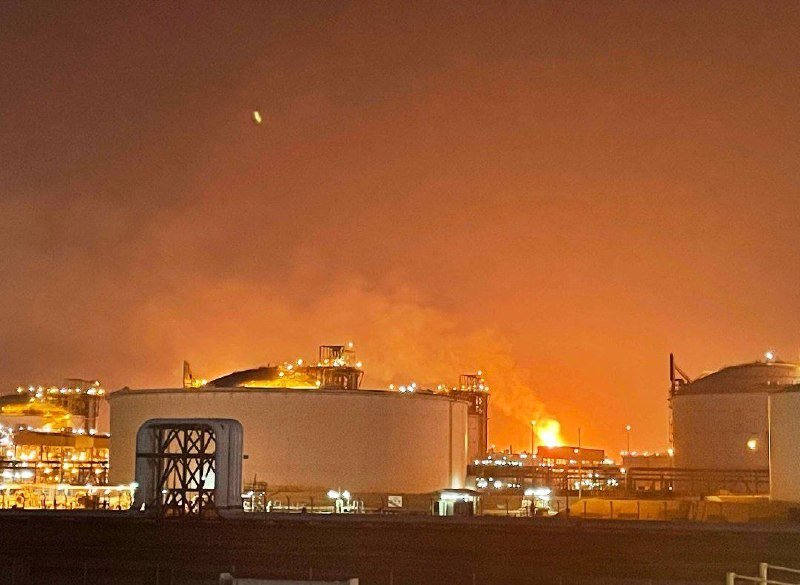 Flames and thick smoke rise from an industrial facility at night, illuminating large storage tanks and surrounding infrastructure with an orange glow.