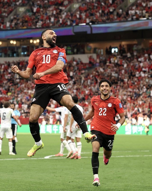 A soccer player wearing a red jersey with number 10 jumps and celebrates while a teammate in red number 22 runs toward him on a pitch in front of a stadium crowd.