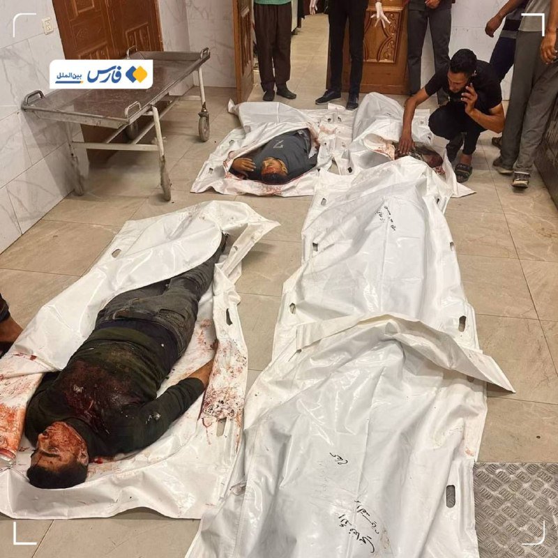 Three bodies lie in white body bags on a tiled floor, with one partially unzipped showing a bloodied man, while a man crouches nearby and others stand around.