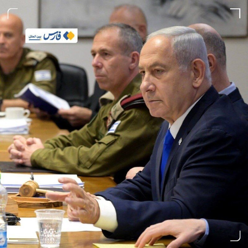 Men in military uniforms and a dark suit sit at a conference table with documents and a water bottle, one gesturing with his hand during what appears to be a meeting.