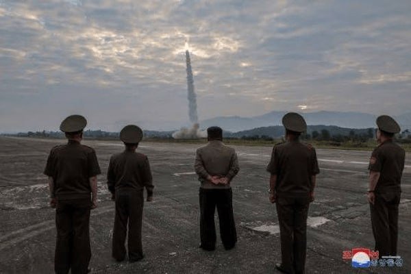Five men in dark military uniforms stand on a flat dirt field, watching a rocket ascend into a cloudy sky in the distance.