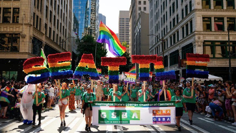Marchers in green shirts carry rainbow floral letters spelling "SEATTLE," a Pride flag, and a GSBA banner down a city street during a parade.