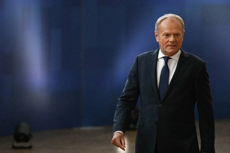 A man in a dark suit and blue tie walks across a stage with a blue backdrop and stage lighting visible.