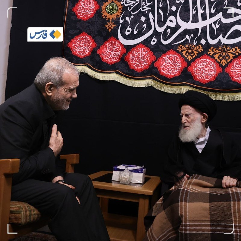 A gray-haired man in a dark suit sits across from a white-bearded cleric in black robes, conversing in a room with Arabic calligraphy banners.