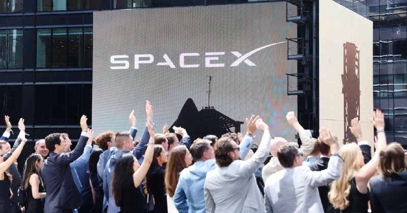A crowd in business attire raises their hands in celebration before a large screen displaying the "SpaceX" logo and a rocket image.
