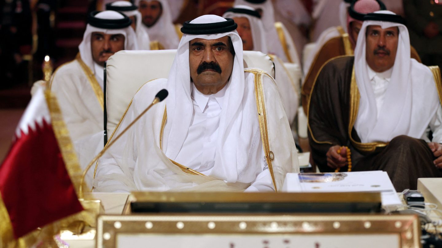 A man in traditional white Gulf attire speaks at a wooden podium bearing an emblem featuring a sailboat, palm trees, and crossed swords.