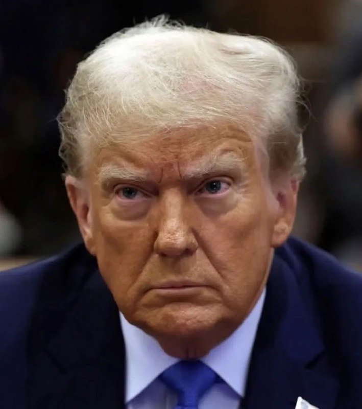 A man with blonde hair wearing a dark suit, white shirt, and blue tie looks forward with a serious expression against a blurred background.