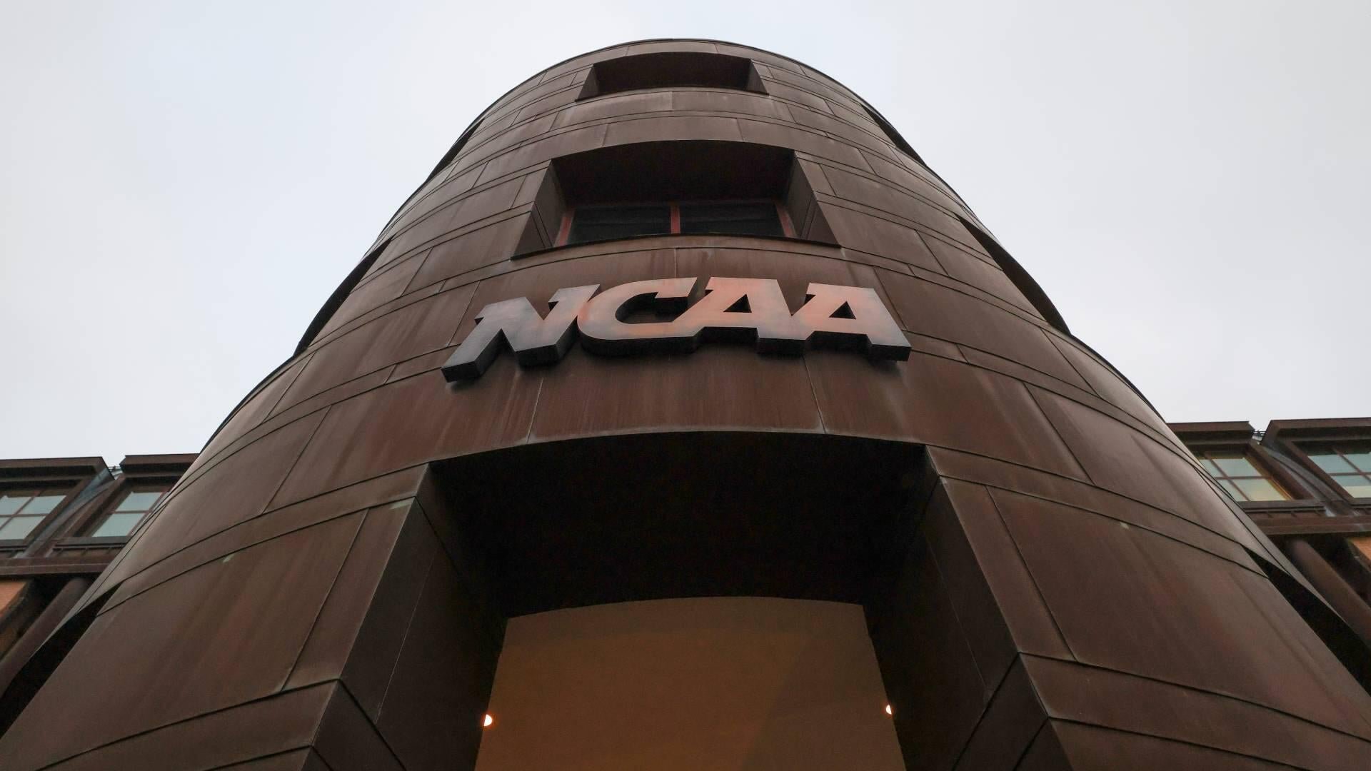 A low-angle photograph shows the NCAA logo displayed on the exterior of a curved, brown-paneled building against an overcast sky.