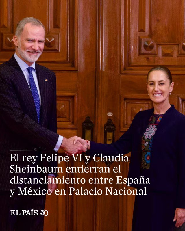 Two formally dressed individuals—a man in a dark suit and a woman in a navy blazer with embroidered blouse—shake hands while smiling in front of large wooden paneled doors.