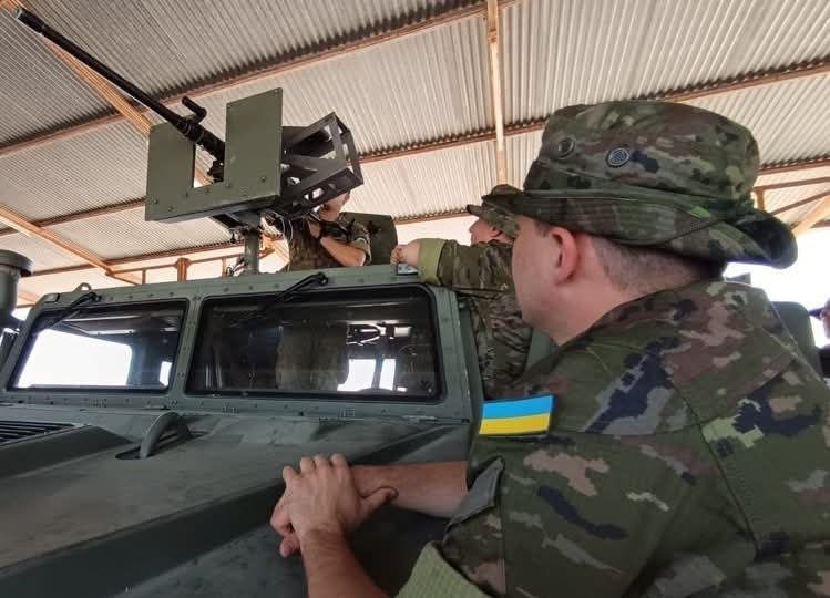 Soldiers in camouflage uniforms examine a mounted weapon on a military vehicle inside a hangar-like structure.