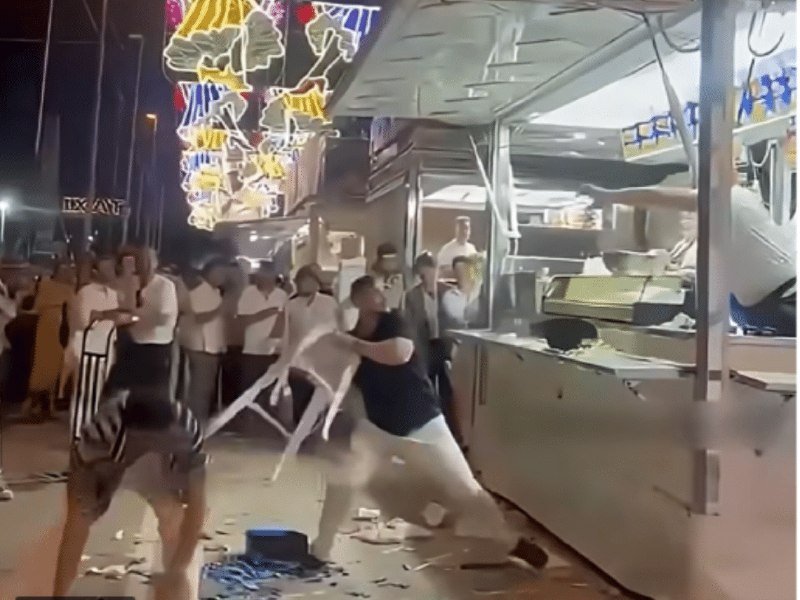 Two people wrestle over a plastic chair at night near a damaged food truck, while onlookers watch under decorative overhead lights and debris litters the ground.