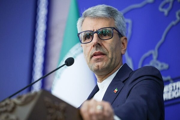 A gray-haired man wearing glasses and a dark suit speaks at a podium with a microphone, backed by an Iranian flag and a blue map display.