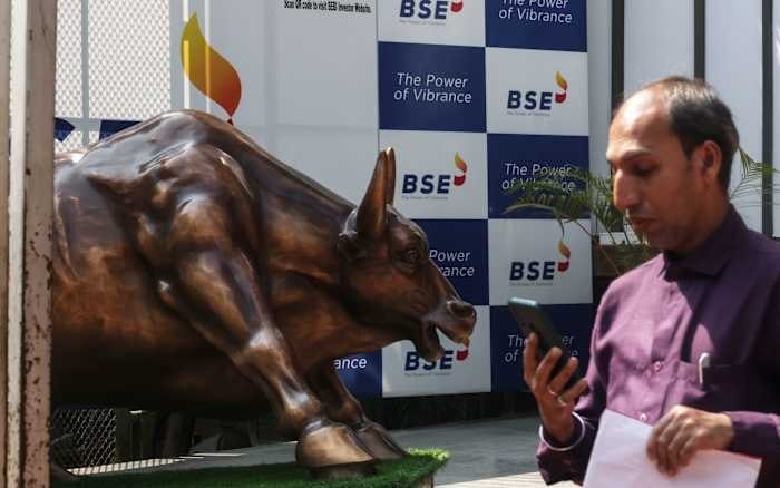 A bronze bull statue stands beside a man in a purple shirt looking at a mobile phone, with a backdrop displaying "BSE" and "The Power of Vibrance" logos.