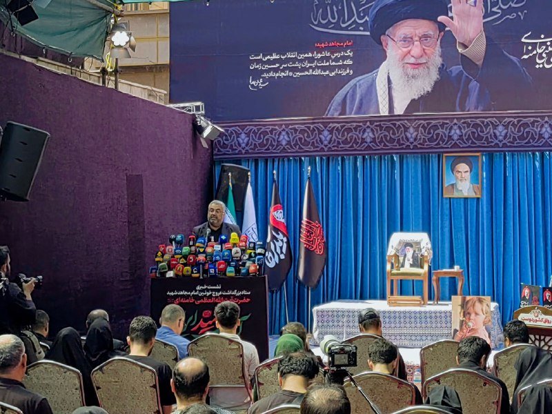 A bearded man speaks at a podium covered with microphones, backed by flags and a large portrait of a cleric.