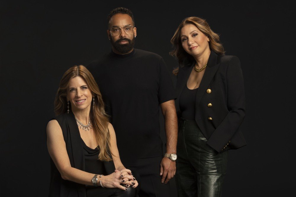 Three people pose against a black background: a seated woman in a black sleeveless top with jewelry, a standing man in a black t-shirt, and a standing woman in a black blazer with gold buttons.