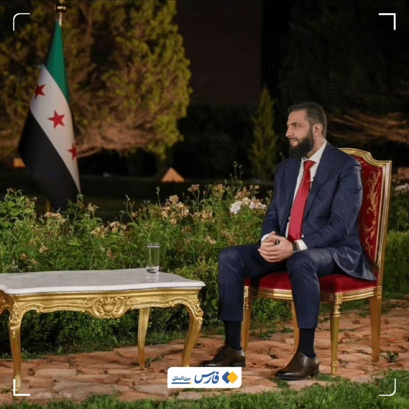 A bearded man in a dark suit and red tie sits in an ornate chair outdoors at night, with a flag and garden visible nearby.