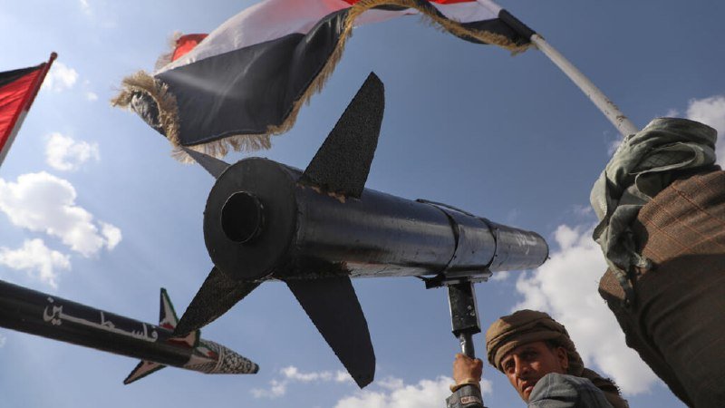 A man holds up a model of a black rocket-like projectile with fins, alongside flags with Arabic text, against a blue sky.