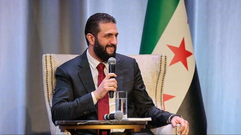 A bearded man in a dark suit and red tie speaks into a microphone while seated beside a flag with red and green stripes and stars.