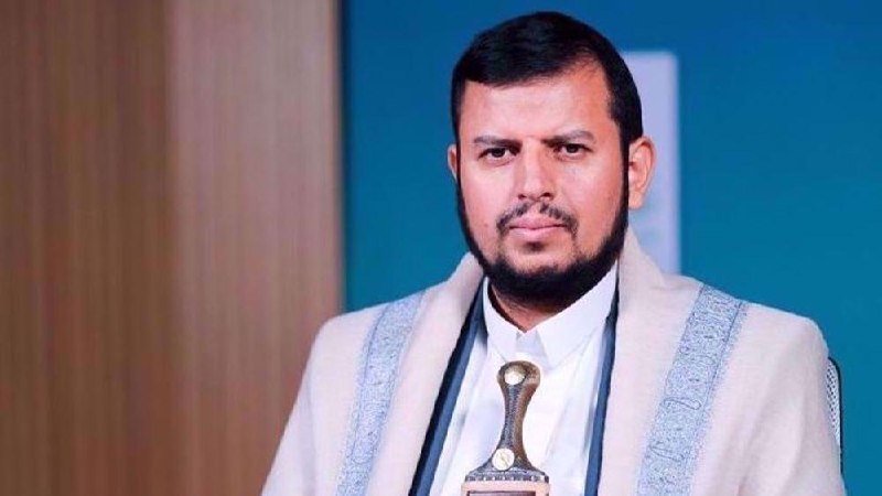 A bearded man in a light-colored traditional robe and white shirt holds a trophy-shaped award against a blue backdrop.
