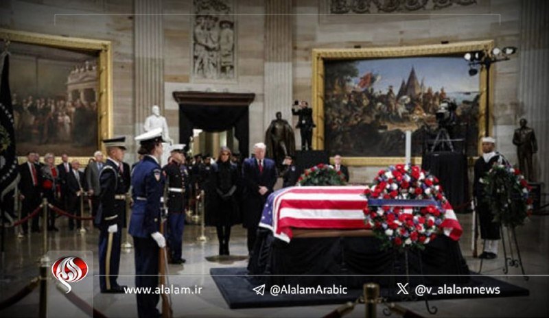 Honor guards stand beside a flag-draped casket topped with red, white, and blue floral arrangements in a grand hall featuring large historical paintings.