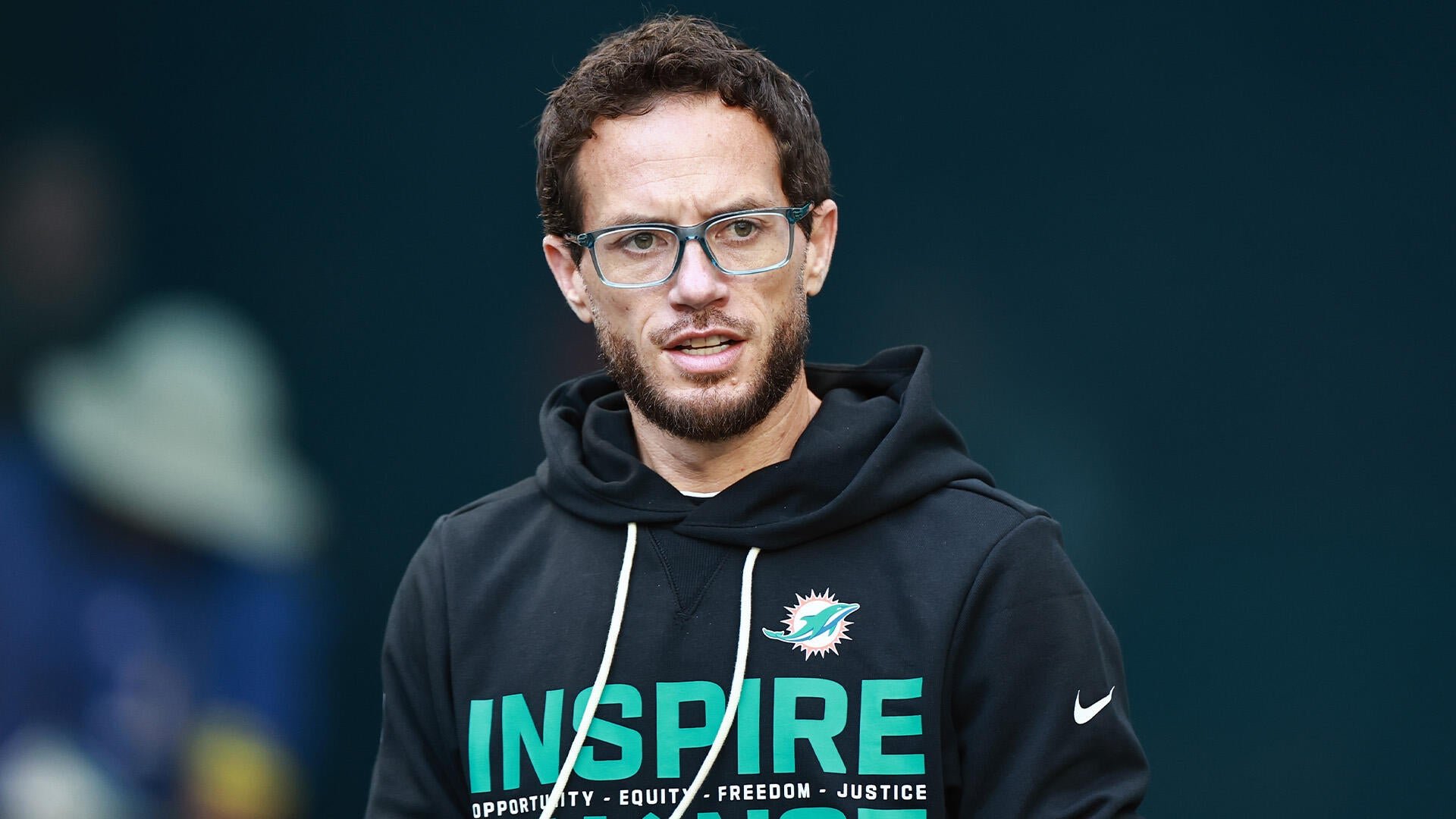 A bearded man wearing glasses and a black Miami Dolphins hoodie with "INSPIRE" text stands against a dark background.