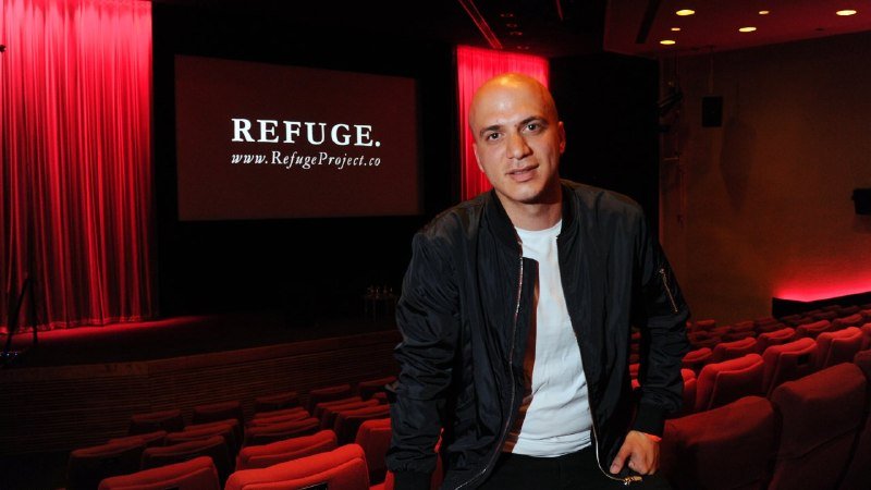 A bald man in a black bomber jacket stands in an auditorium, with a screen behind him displaying "REFUGE." and "www.RefugeProject.co" beside red curtains and empty theater seats.