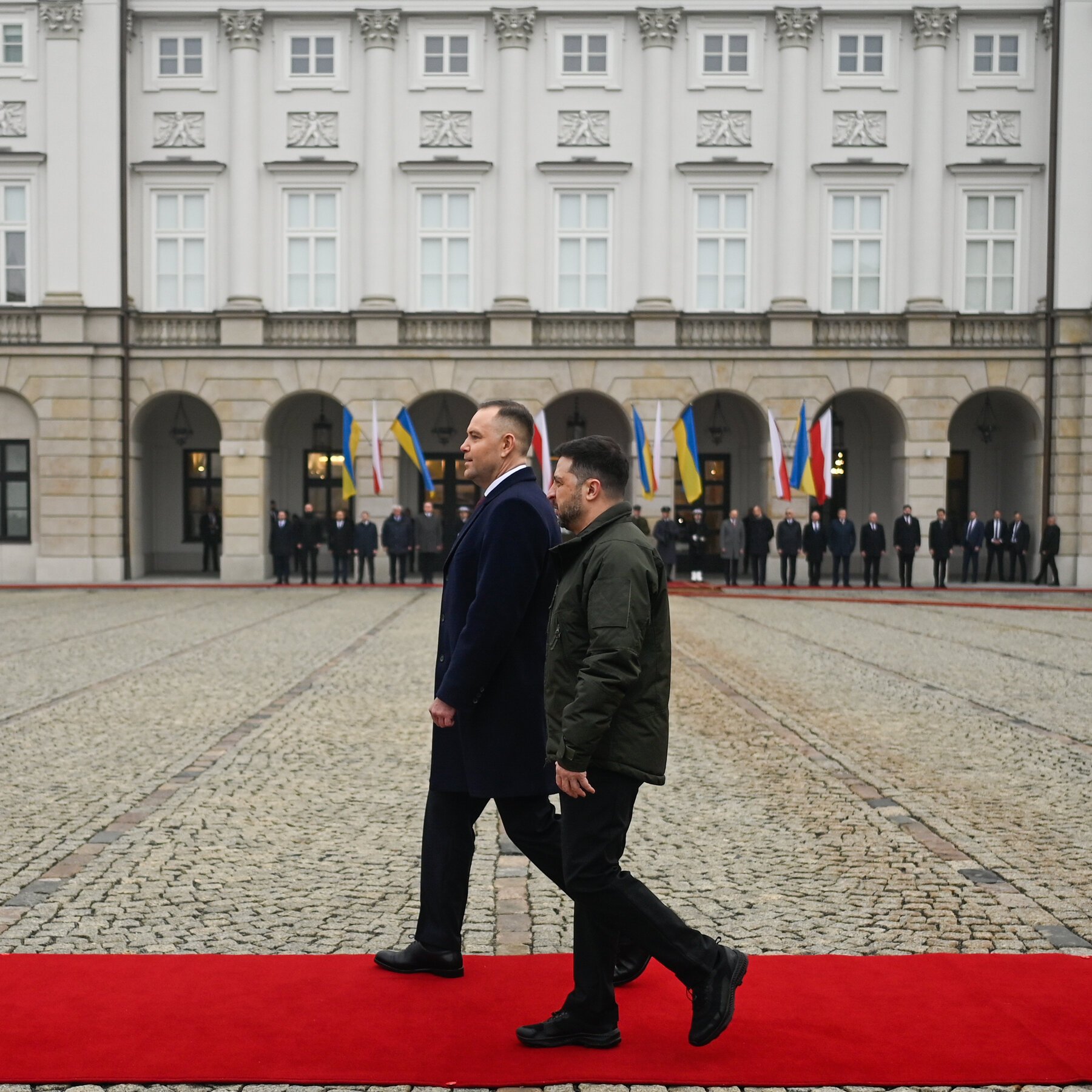Two men in dark coats walk along a red carpet in a cobblestone courtyard before a grand classical building adorned with multiple flags, while a line of officials stands under the arched colonnade.