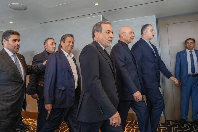 Seven men in formal suits walk together through a modern, well-lit interior hallway with patterned carpet and neutral walls.