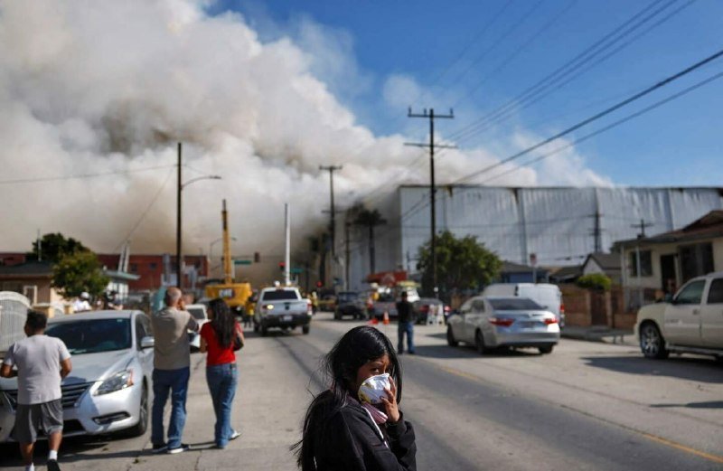 A young girl covers her face with a cloth while a large plume of smoke rises from a structure fire down a residential street, with onlookers and parked cars nearby.
